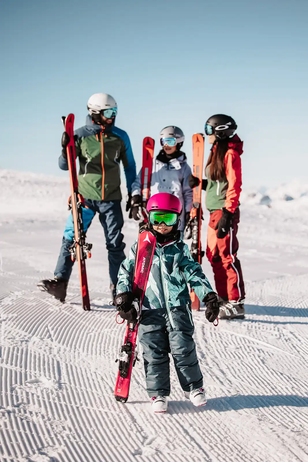 Skifamilie auf der präparierten Skipiste – im Vordergrund ein Kind mit pinkem Helm und Skiern in der Hand, im Hintergrund drei Erwachsene in Skibekleidung mit Ausrüstung, vor strahlend blauem Himmel.
