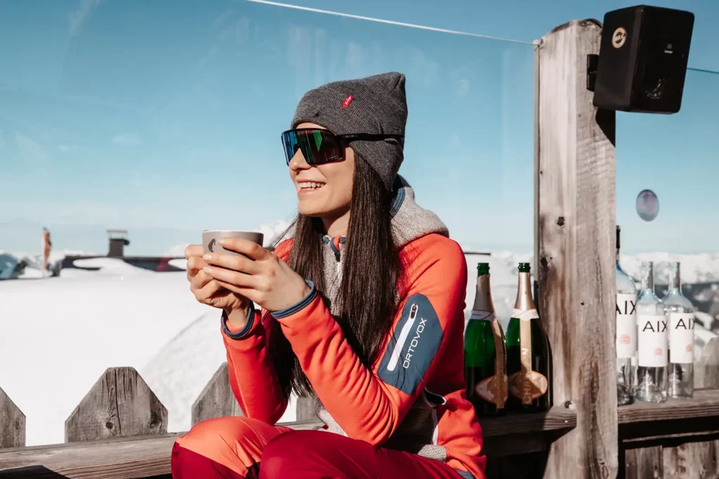 Frau in winterlicher Sportkleidung genießt mit einer Tasse Heißgetränk die Sonne auf einer Holzbank vor verschneiter Bergkulisse – entspannter Moment auf der Hütte mit Weitblick.