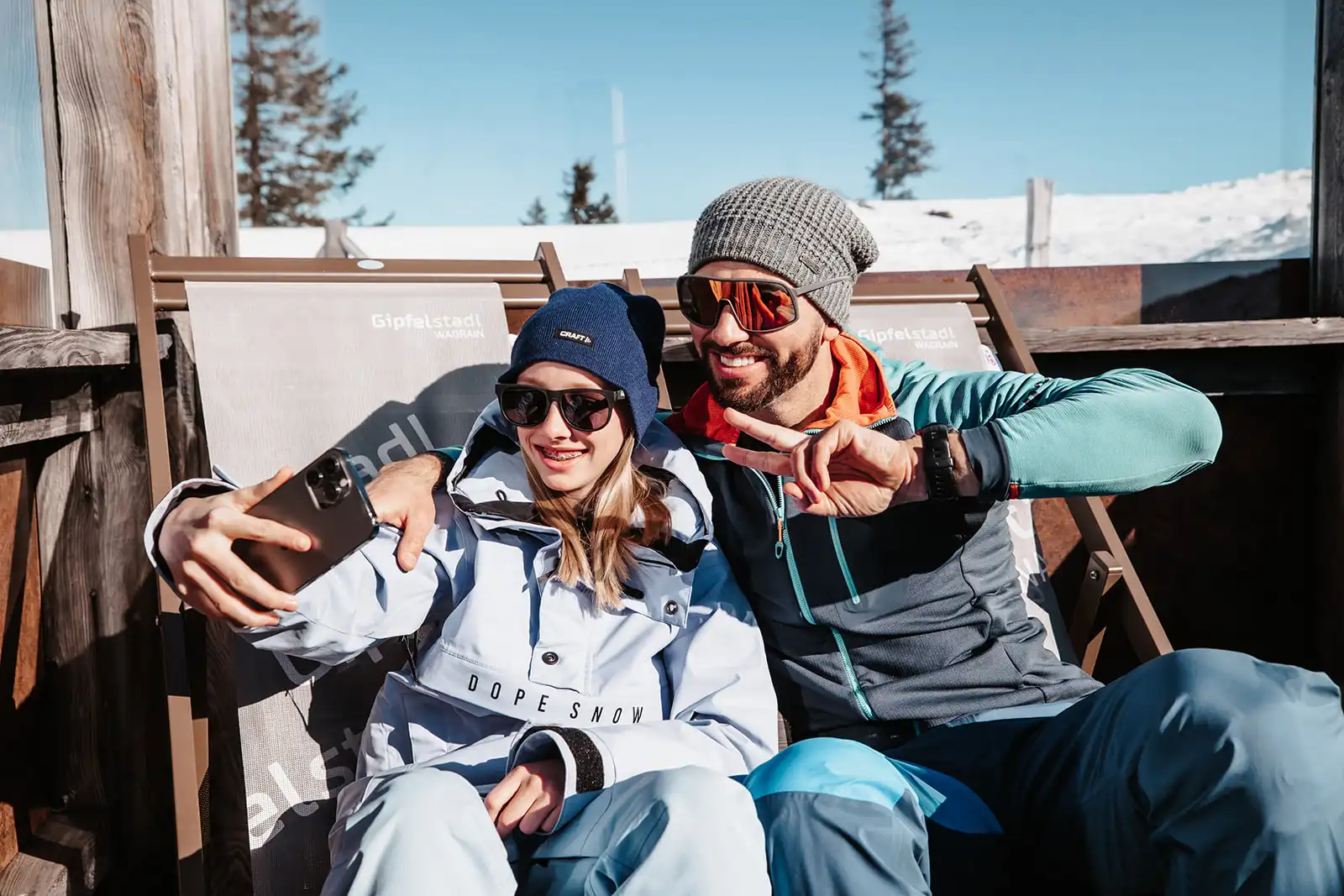 Vater und Tochter in Winterkleidung entspannen in Liegestühlen vor einer Berghütte und machen gemeinsam ein fröhliches Selfie – strahlender Sonnenschein und verschneite Kulisse im Hintergrund.