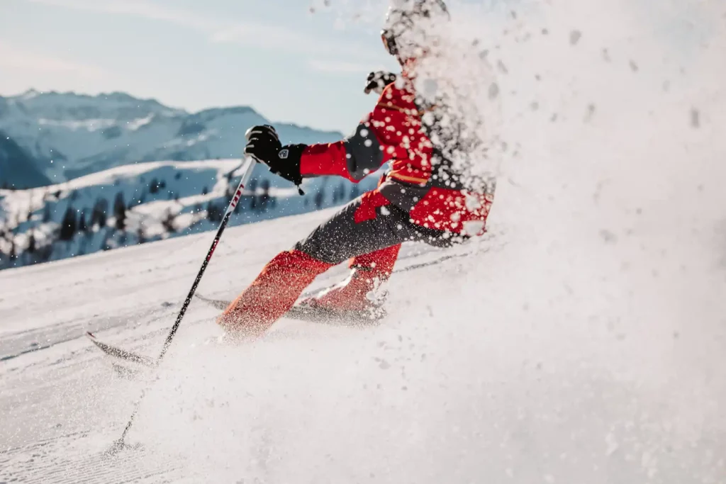 Dynamisk billede af en skiløber i rødt skitøj, der bevæger sig i høj fart gennem frisk puddersne - sneen hvirvler op foran en imponerende bjergkulisse.