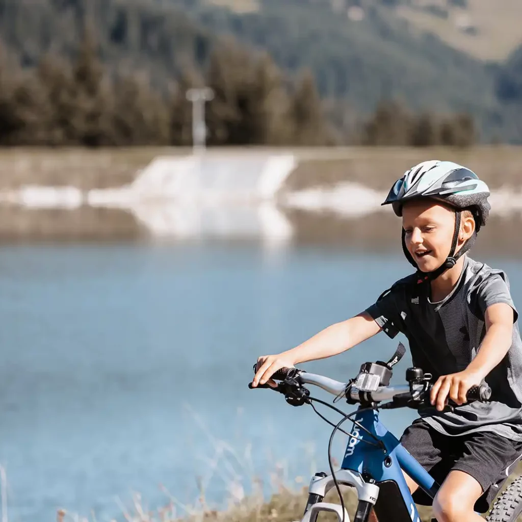 Hofgut©Elisabeth_Fotografie-207 A boy in a grey shirt and bicycle helmet rides along the shore of a lake on a blue mountain bike, laughing.