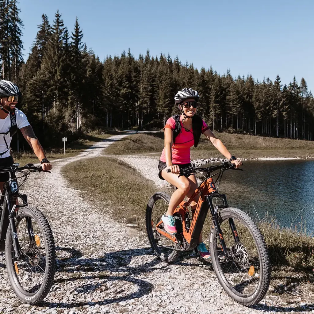 Hofgut©Elisabeth_Fotografie-213 A man and a woman wearing cycle helmets ride along a winding gravel track on mountain bikes, with a dense forest and lake in the background.