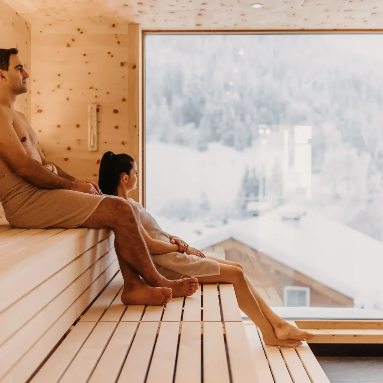 Wohlfühlmoment mit Panoramablick: Ein Paar genießt die wohltuende Wärme der Skysauna im Hofgut Wagrain mit Blick auf die verschneiten Dächer der alpinen Winterlandschaft.