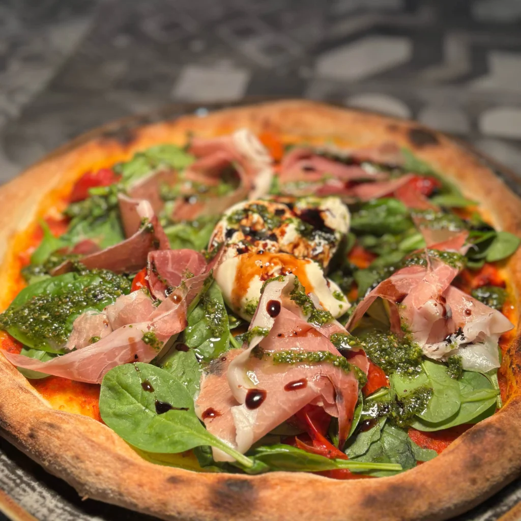 Pizza Burrata Frisch gebackene Pizza mit knusprigem Rand, belegt mit Prosciutto, frischem Spinat, Burrata, grünem Pesto und einem Hauch Balsamico-Glasur.