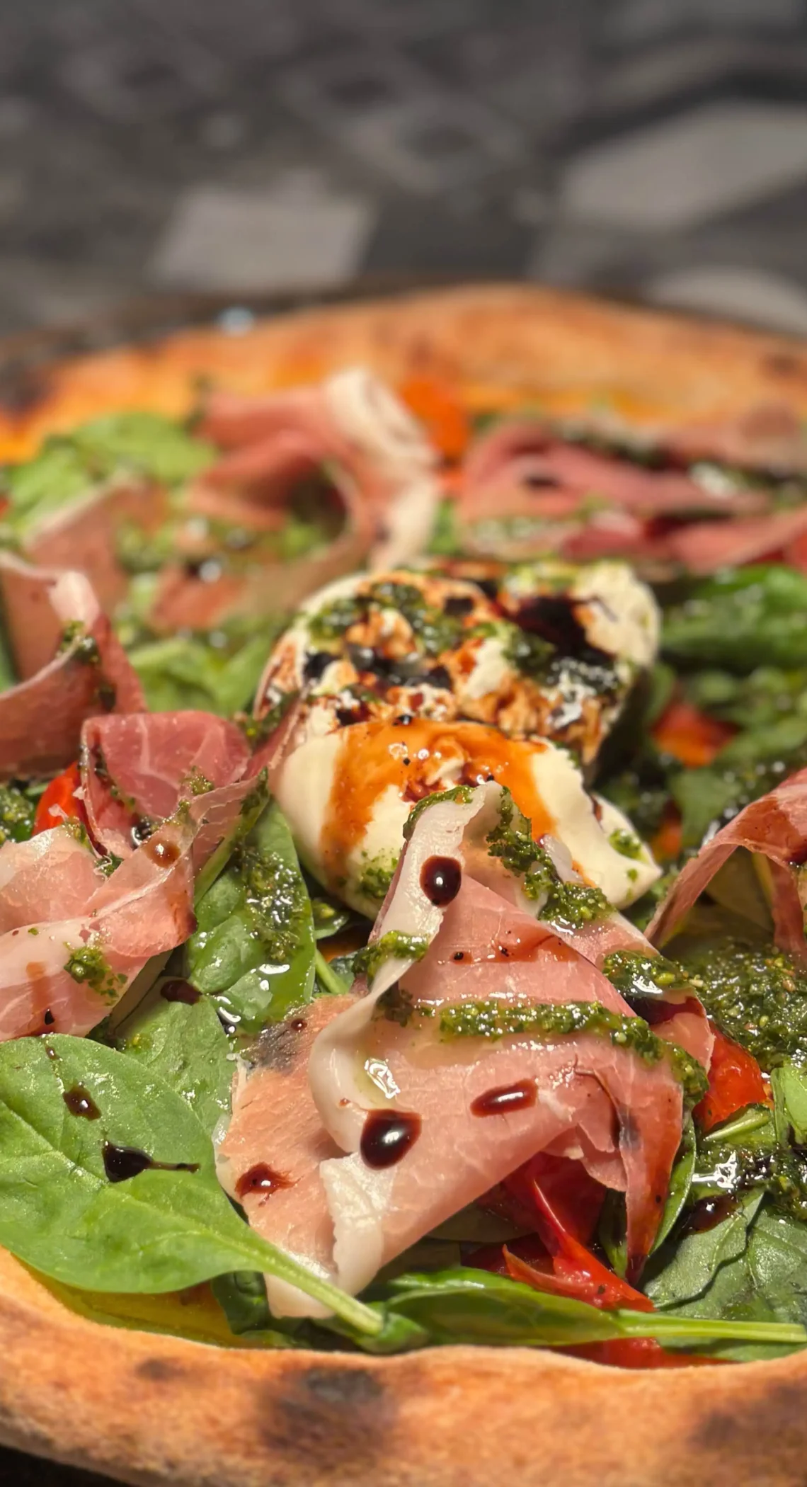 Frisch gebackene Pizza mit knusprigem Rand, belegt mit Prosciutto, frischem Spinat, Burrata, grünem Pesto und einem Hauch Balsamico-Glasur.