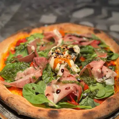 Frisch gebackene Pizza mit knusprigem Rand, belegt mit Prosciutto, frischem Spinat, Burrata, grünem Pesto und einem Hauch Balsamico-Glasur.