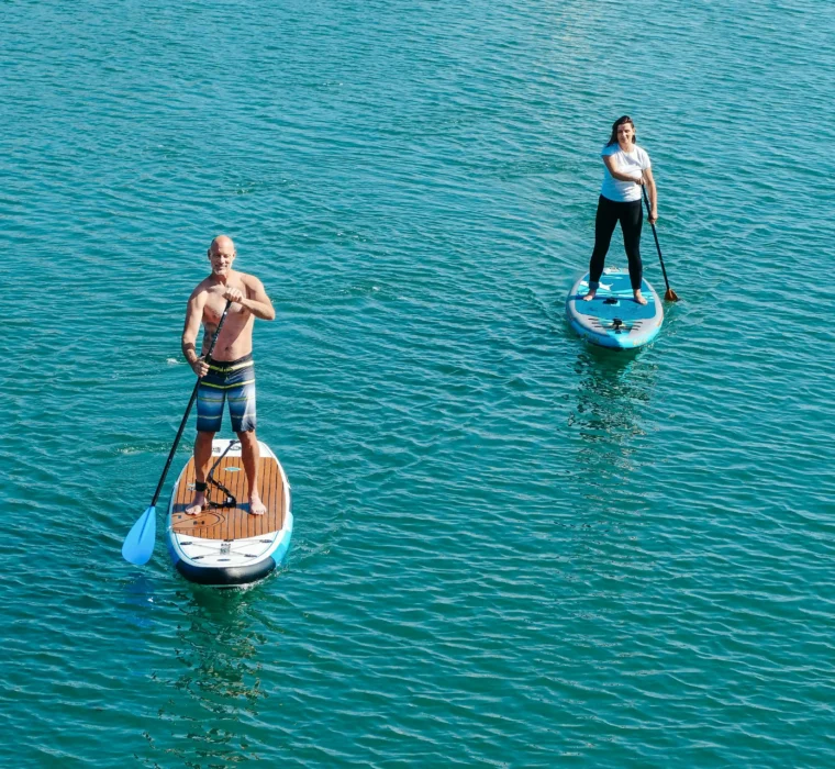 Zwei Personen stehen auf ihren SUP-Boards auf ruhigem, blauem Wasser und paddeln entspannt nebeneinander.