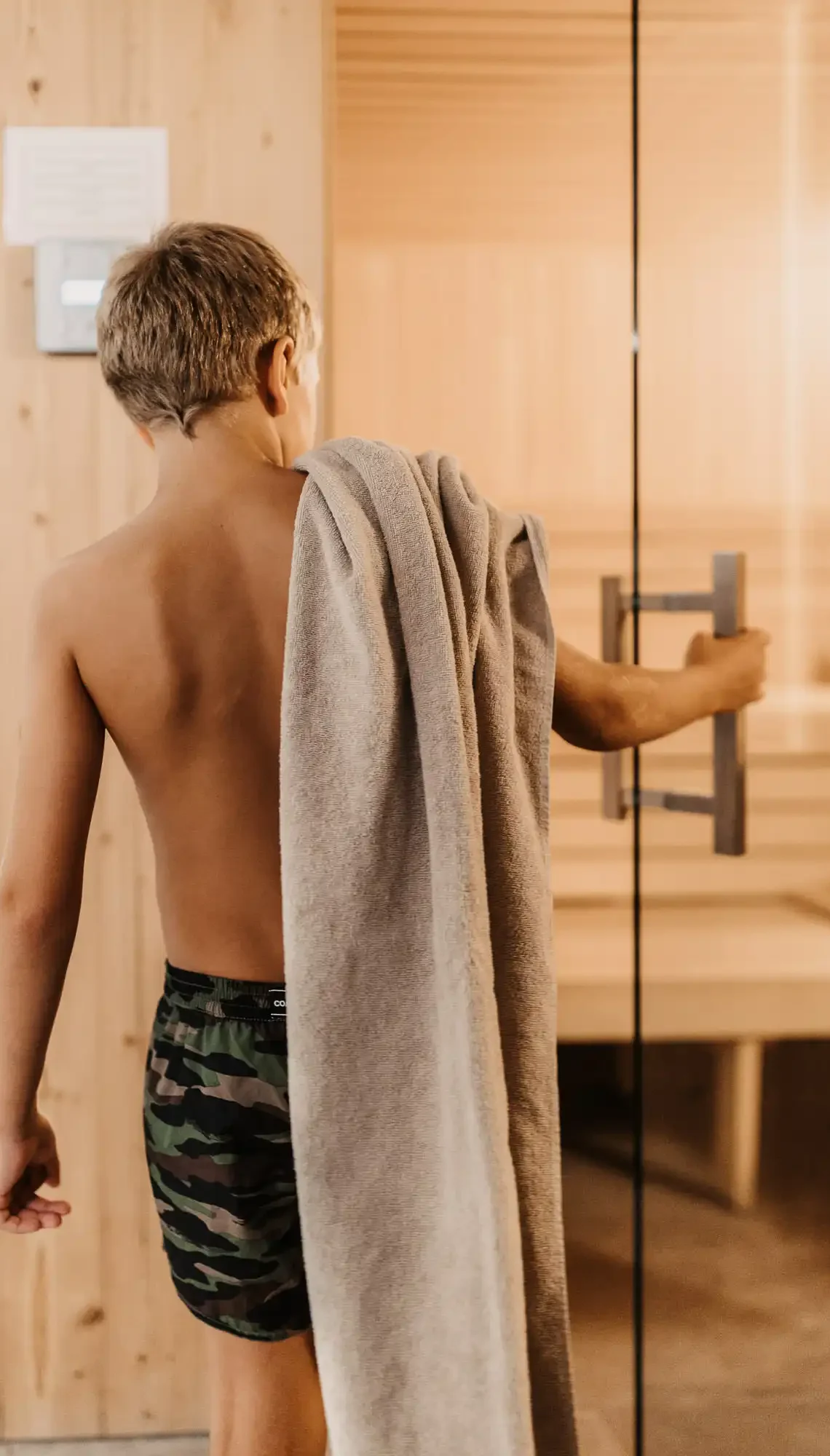 Ein Kind geht mit einem Handtuch über der Schulter in eine helle Sauna und öffnet die Glastür – ein Moment entspannter Familienwellness.