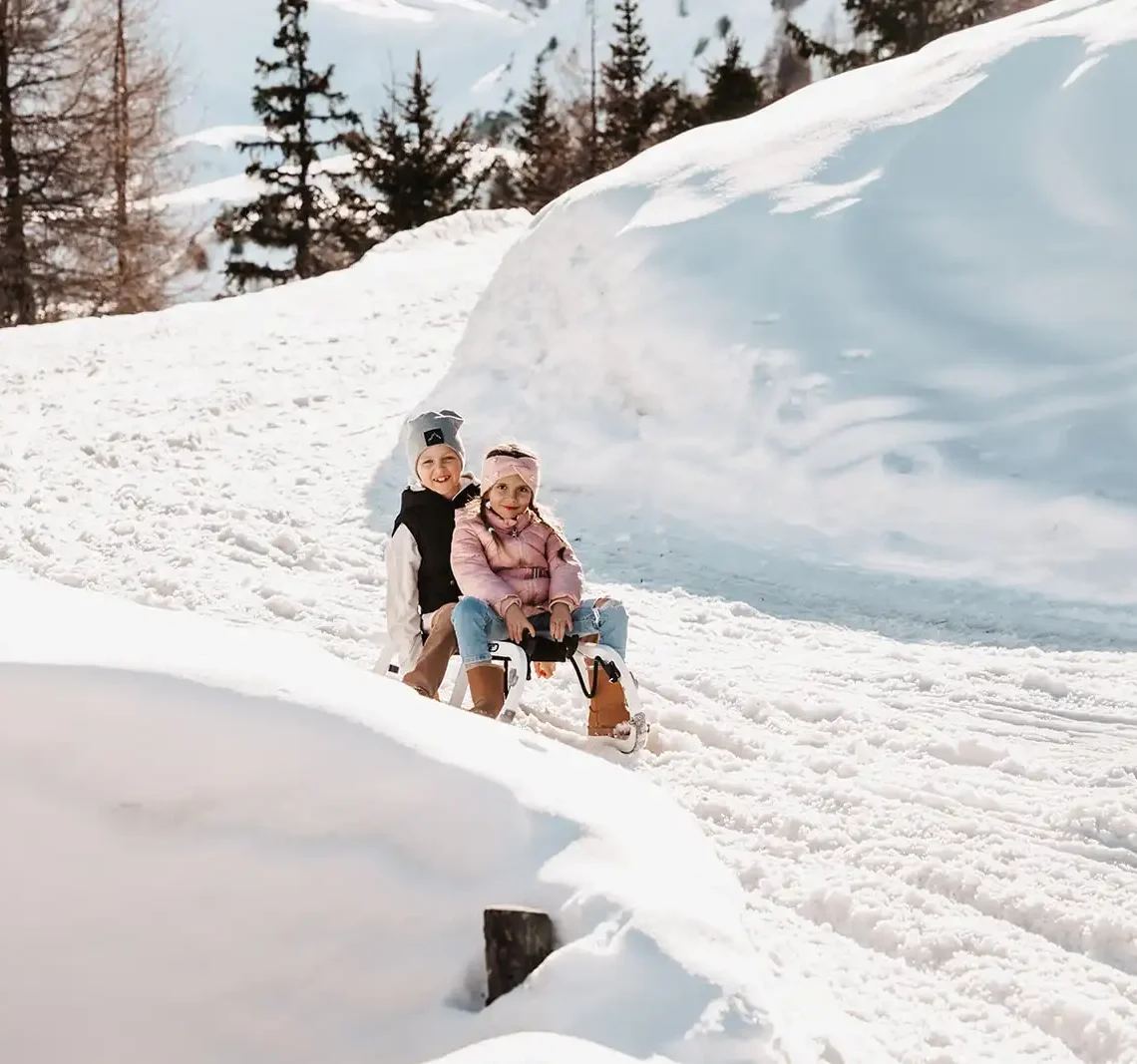 Zwei Kinder auf einem Schlitten, lachend, in Winterkleidung auf verschneitem Hang.