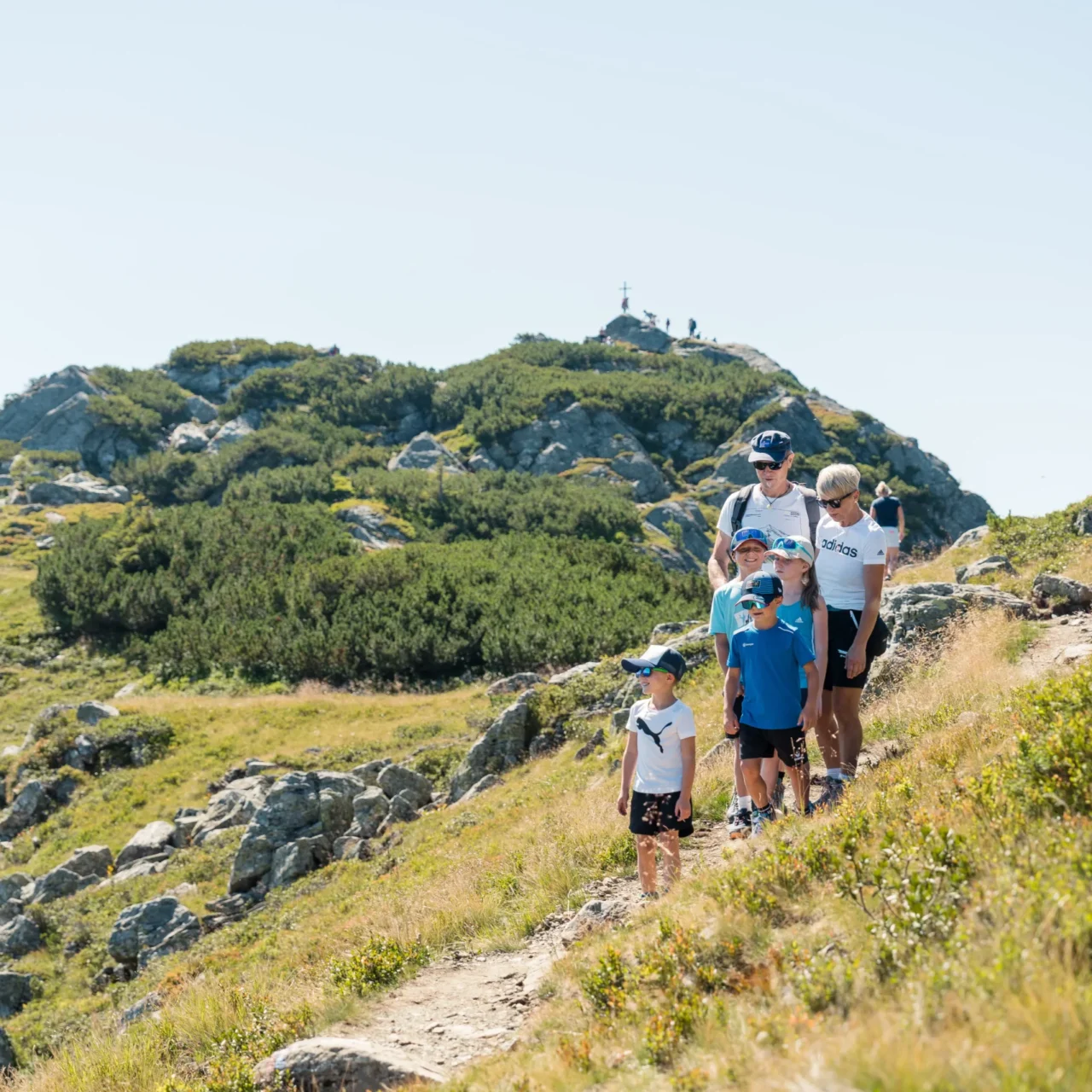 Familie mit Kindern wandert auf einem sonnigen Bergpfad durch die alpine Salzburger Landschaft, umgeben von Felsen, Almwiesen und einem Gipfel im Hintergrund.