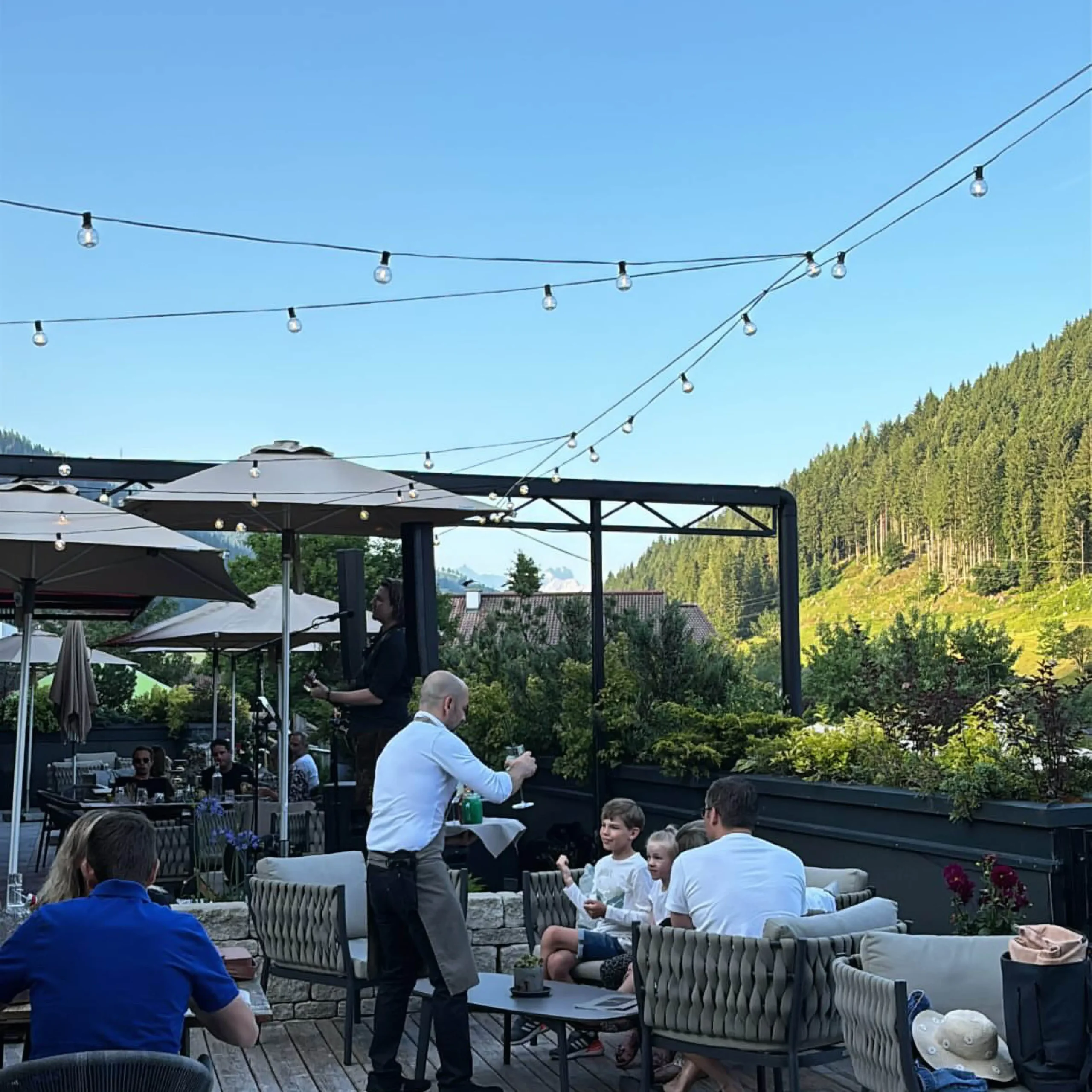 Sommerliche Terrasse im Restaurant Vamoos im Hofgut Wagrain mit Gästen, Lichterketten und Blick auf die umliegenden Berge.