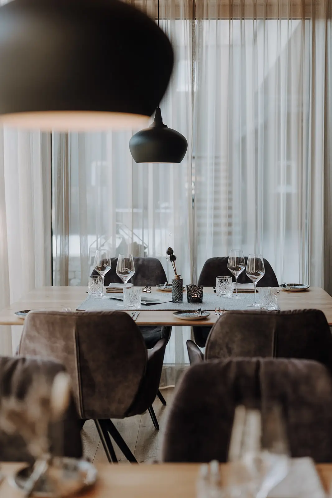 Stilvoll gedeckter Tisch im Restaurant Vamoos im Hofgut Wagrain mit Weingläsern, gepolsterten Stühlen und moderner Pendelleuchte vor großen, lichtdurchlässigen Vorhängen.