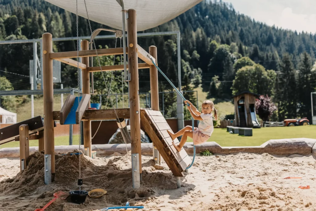 Alt-Text: Kleines Kind spielt auf einem Holz-Klettergerüst mit Seil in einer Sandkiste am Outdoor-Spielplatz des Hofgut Wagrain, vor alpiner Bergkulisse.
