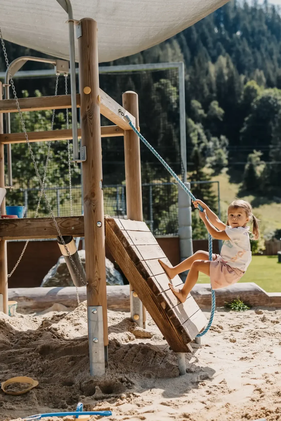 Kleines Kind spielt auf einem Holz-Klettergerüst mit Seil in einer Sandkiste am Outdoor-Spielplatz des Hofgut Wagrain, vor alpiner Bergkulisse.