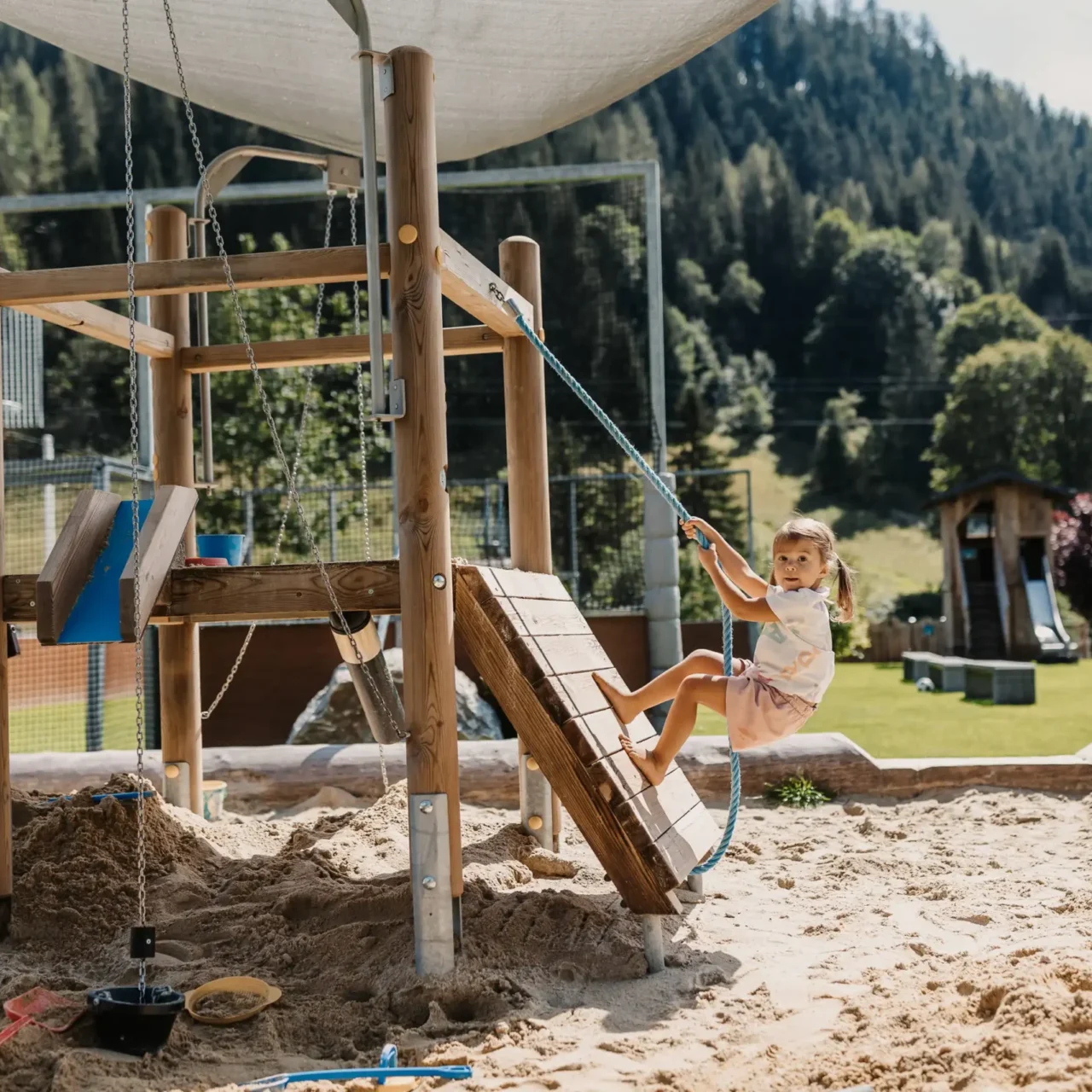 Kleines Kind spielt auf einem Holz-Klettergerüst mit Seil in einer Sandkiste am Outdoor-Spielplatz des Hofgut Wagrain, vor alpiner Bergkulisse.