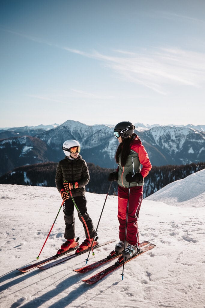 Mutter und Kind in Skiausrüstung lachen gemeinsam auf der Piste vor winterlicher Bergkulisse.