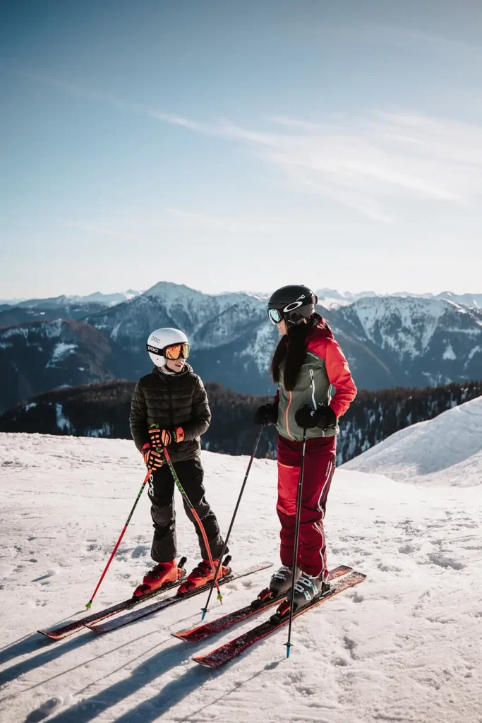 Mother and child in ski gear laughing together on the slopes in front of a wintry mountain landscape.